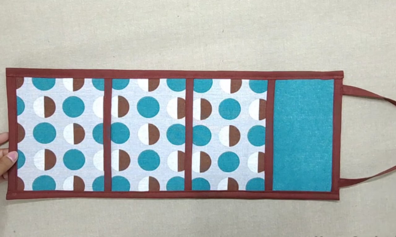 A handmade fabric wall organizer with four vertical pockets. The organizer is made of blue, white, and brown patterned fabric, framed with a dark red border. The leftmost pocket is being held open by a hand. The rightmost pocket is solid teal and slightly larger, with a strap attached for hanging.