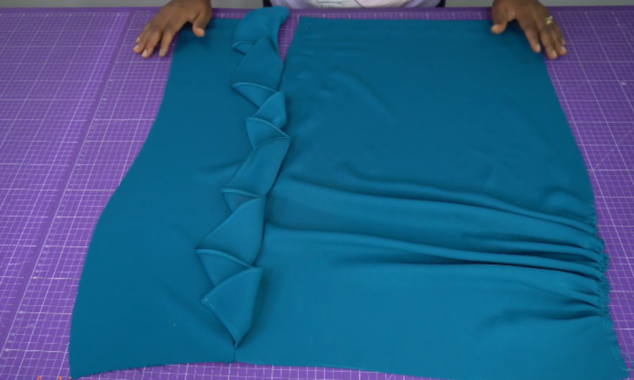 A person arranging teal blue fabric pieces on a purple cutting mat. The left piece features decorative folded petal-like details down the seam, and the right piece is gathered on one side, likely as part of assembling a pencil skirt with unique textural design.