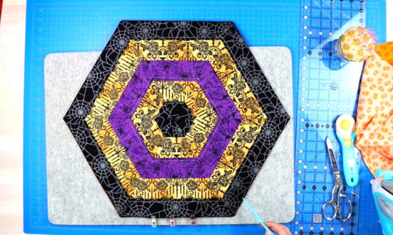 A completed hexagon table topper made with Halloween-themed fabric strips in black, gold, and purple. The fabric has patterns like spider webs, skulls, and roses. The topper is placed on a gray felt pad over a blue cutting mat. Quilting tools such as a ruler, rotary cutter, scissors, and fabric clips are visible on the side.