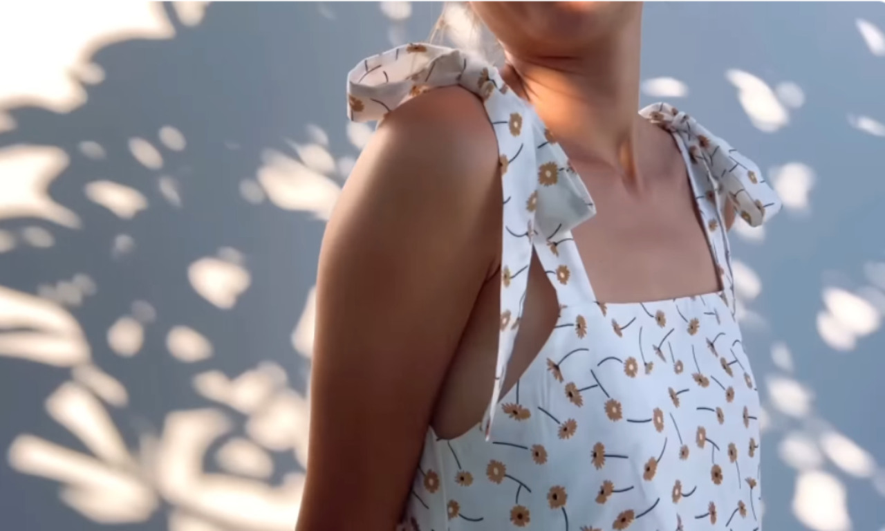 Woman wearing a sleeveless white dress with a yellow daisy print, featuring tied shoulder straps and a square neckline, standing in dappled sunlight.