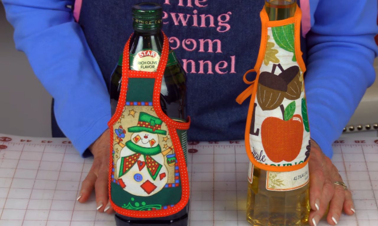 Two completed small aprons are displayed on wine or olive oil bottles. The left bottle has a green apron with a festive snowman design, while the right bottle has an autumn-themed apron with acorns and an apple.