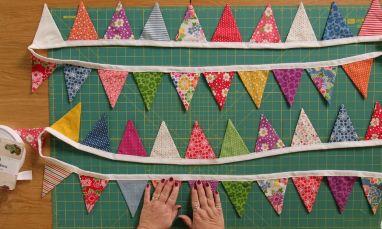A green cutting mat displays several colorful fabric bunting strings made of triangle-shaped flags sewn onto white bias tape. The triangles are in various colors and patterns, including polka dots, stripes, and florals. A pair of hands with red-painted nails and rings is seen arranging the bunting pieces at the bottom.