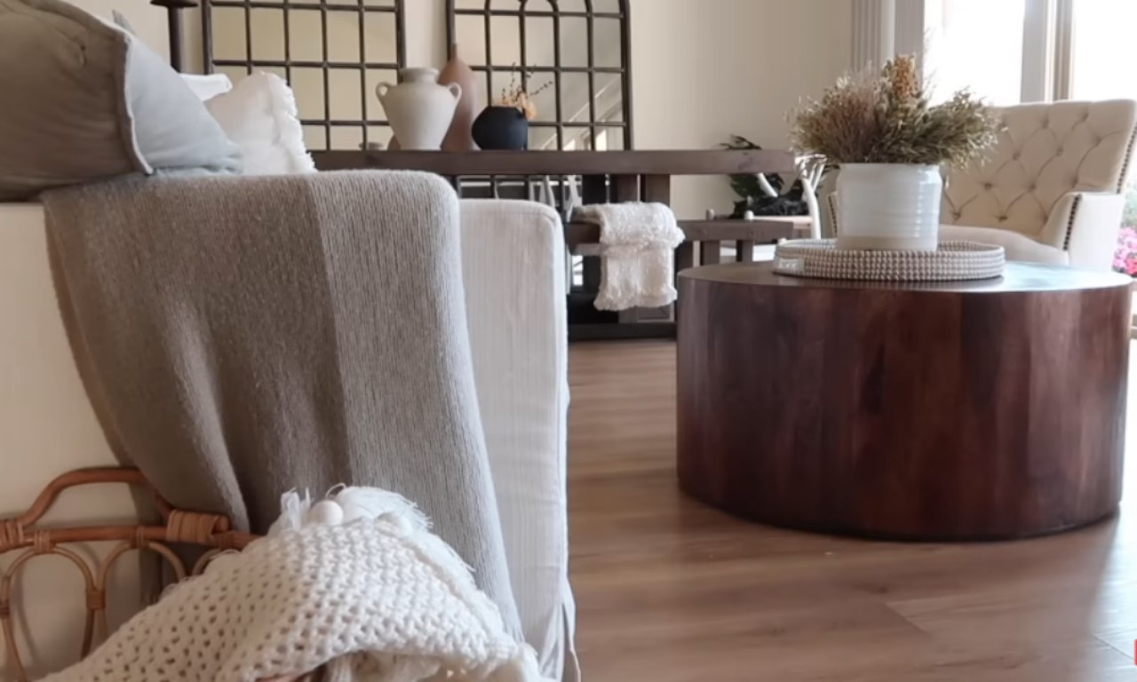 A living room with a neutral-colored sofa covered in a brown knitted throw blanket. In the center is a round, dark wood coffee table with a white pot of dried flowers and a decorative tray on top. In the background, there's a dining table and a tufted armchair.