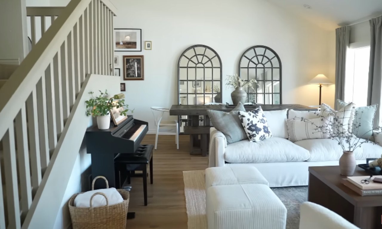 A bright, open-plan living and dining room. To the left is a staircase with light wood banisters and a black upright piano underneath. The living area features a large, slipcovered white sofa with neutral and floral pillows, two white tufted ottomans, and a dark coffee table. The dining area behind the sofa has a dark wood table flanked by two large, arched black-framed mirrors. A basket with rolled-up white blankets is on the floor by the piano. The walls are white and the flooring is light wood.