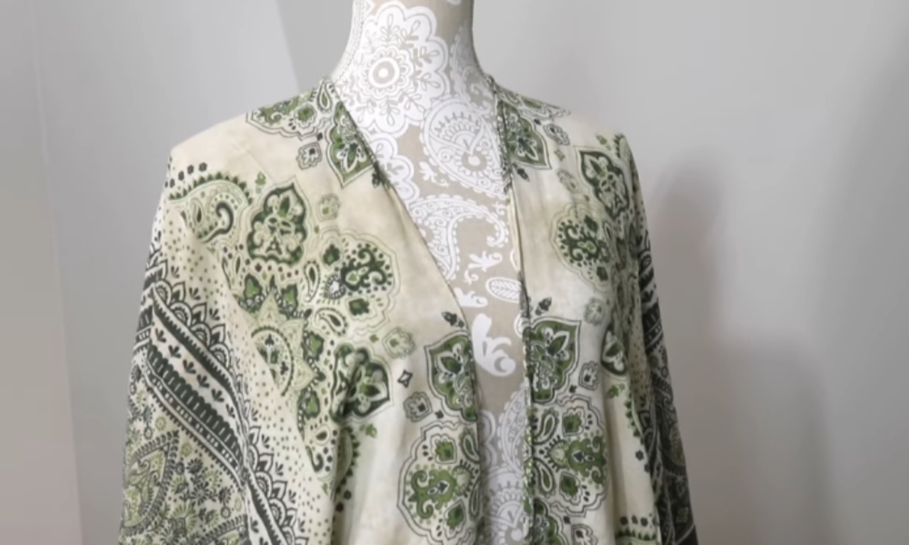 A green and white patterned beach cover-up displayed on a mannequin. The fabric features a paisley and floral motif, and the garment has an open front with flowing sleeves. The mannequin itself has a decorative floral pattern.
