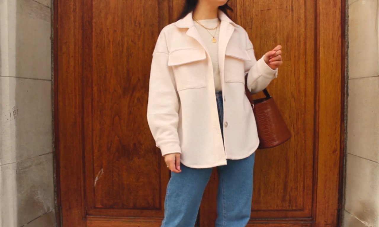 A person wearing a light beige oversized shacket, blue jeans, and carrying a brown leather bucket bag, standing in front of a wooden door. The shacket features flap pockets and a relaxed fit.