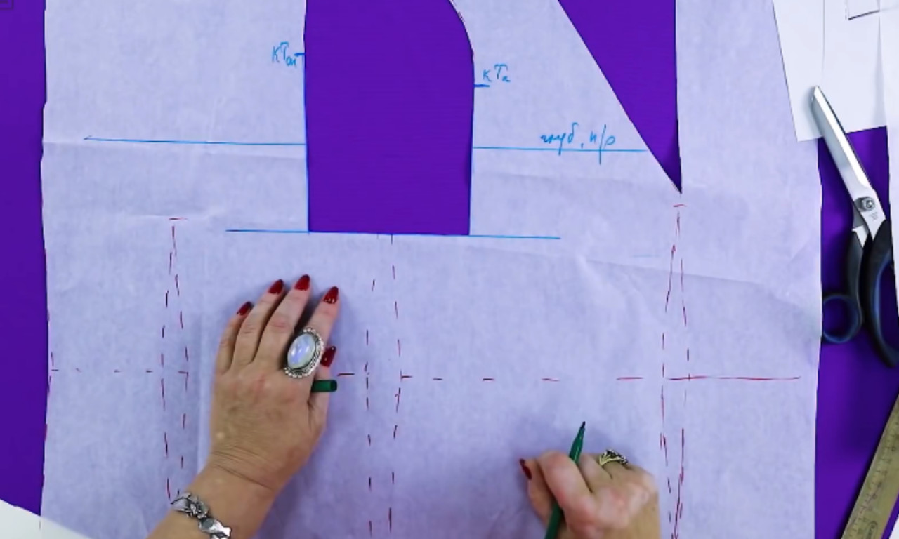 A person is working on a sewing pattern using tracing paper, drawing dotted red lines to mark seam allowances or cutting guides. The person is using a green pencil and is wearing large rings. Scissors and a ruler are placed nearby on a purple surface.