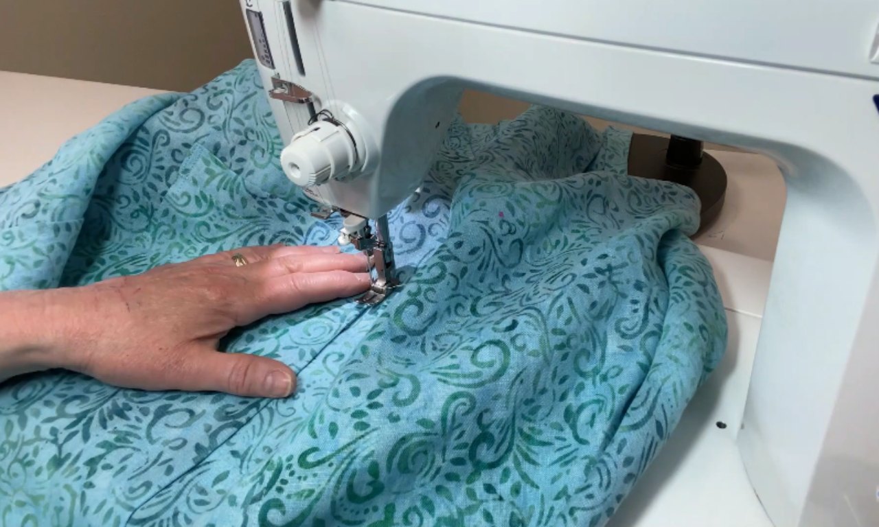 A close-up of hands guiding blue patterned fabric through a white sewing machine, stitching a seam. The fabric features swirly, leafy designs in shades of green and teal.
