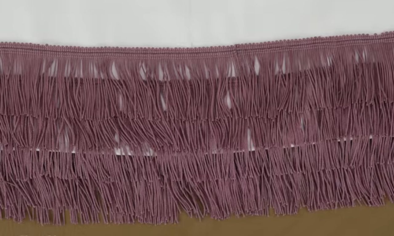 Finished view of a layered fringe design, showing several horizontal rows of mauve fringe sewn onto white fabric, creating a textured, tiered effect.