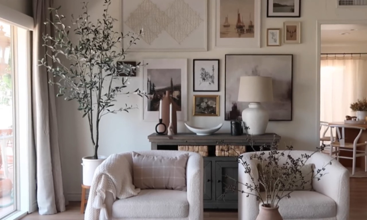 A neutral-toned living room with two comfortable, textured off-white armchairs in front of a gallery wall. The wall features a mix of framed art, mostly in muted tones and varying sizes. Between the chairs is a dark grey console table decorated with a large ceramic lamp, candles, and decorative bowls. A tall, faux olive tree stands to the left.