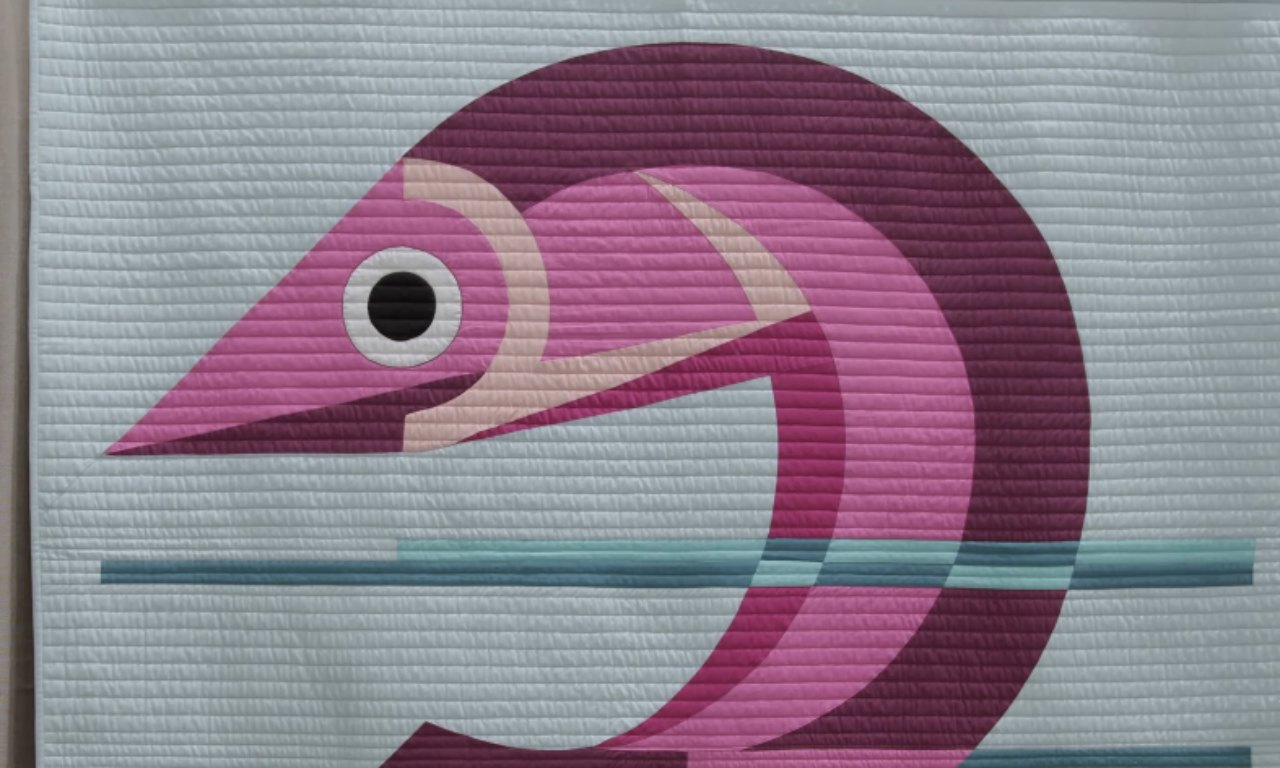 A whimsical quilt design of a stylized pink and maroon fish with a bold circular eye, set against a light blue background with horizontal quilting that mimics water ripples.