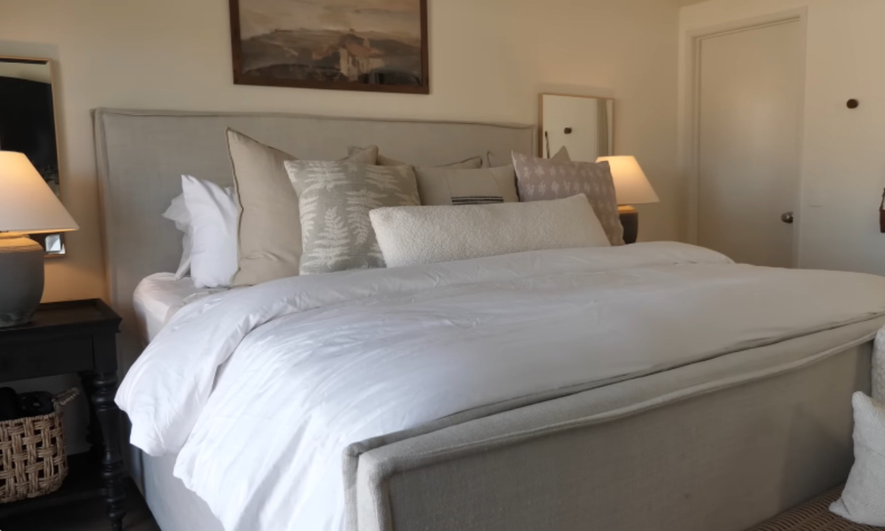 A bedroom scene showing a large, linen-upholstered bed frame with a padded headboard. The bedding is primarily white, layered with several throw pillows in neutral shades of white, cream, and subtle patterns. A wooden nightstand is visible on the left, topped with a lamp.