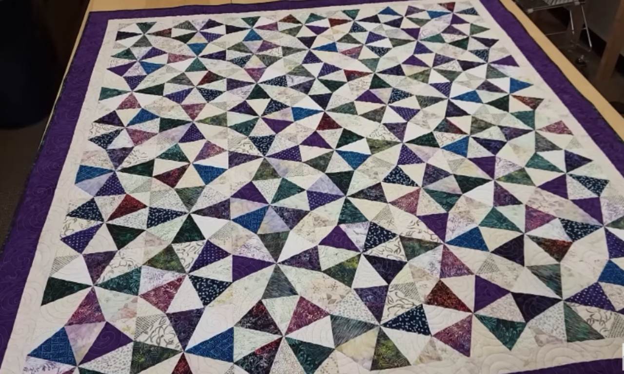 A finished quilt composed of multiple colorful pinwheel-style blocks made from triangle-shaped fabrics arranged in circular patterns, bordered with a rich purple edge.