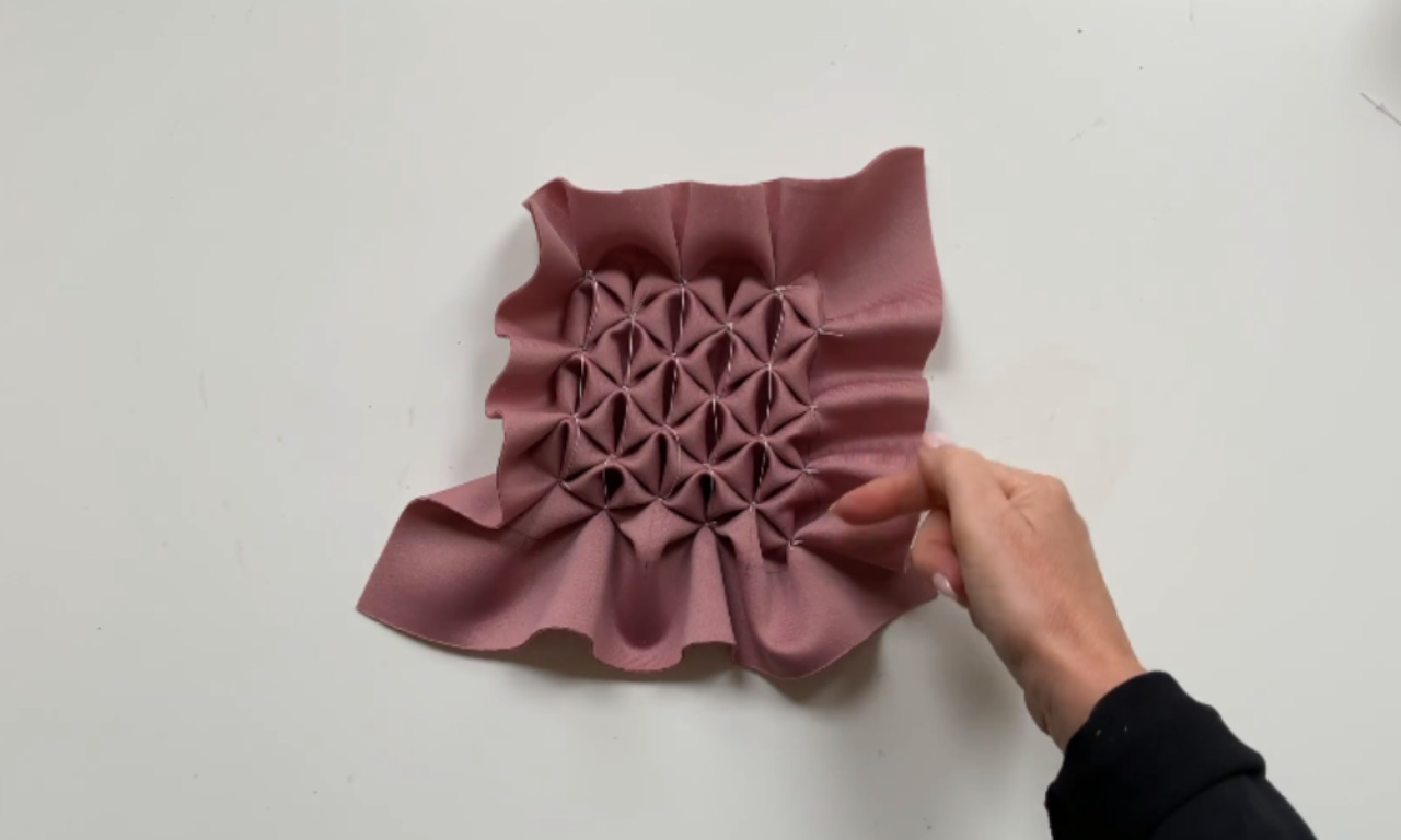 A hand gently holds the edge of a piece of dusty rose-colored fabric that has been hand-smocked into a repeating geometric floral pattern, creating a gathered and textured effect in the center.