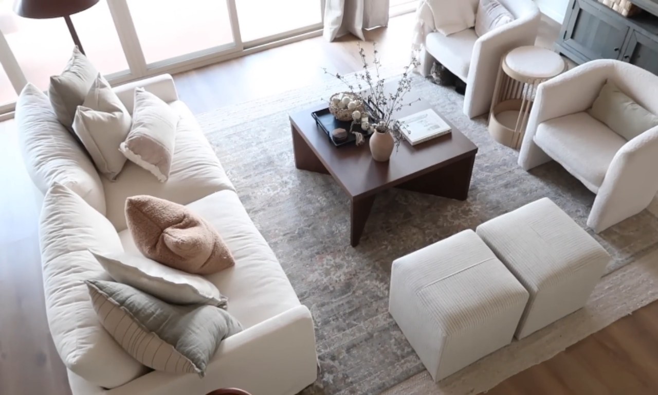 An overhead view of a neutral, contemporary living room. The room features a large white modular sofa piled with neutral throw pillows, a dark brown triangular or uniquely shaped coffee table with minimal styling, and two rounded, off-white armchairs. A light-colored patterned area rug covers the hardwood floor.