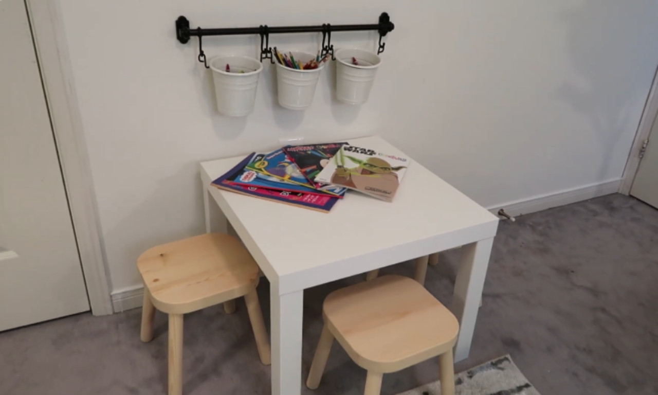 A small, square white table with two light wooden stools in a play area. On the table are several children's books and coloring books, including one featuring Yoda. Above the table, a black wall rail holds three white metal buckets filled with crayons and colored pencils. The floor is covered in a light gray carpet.