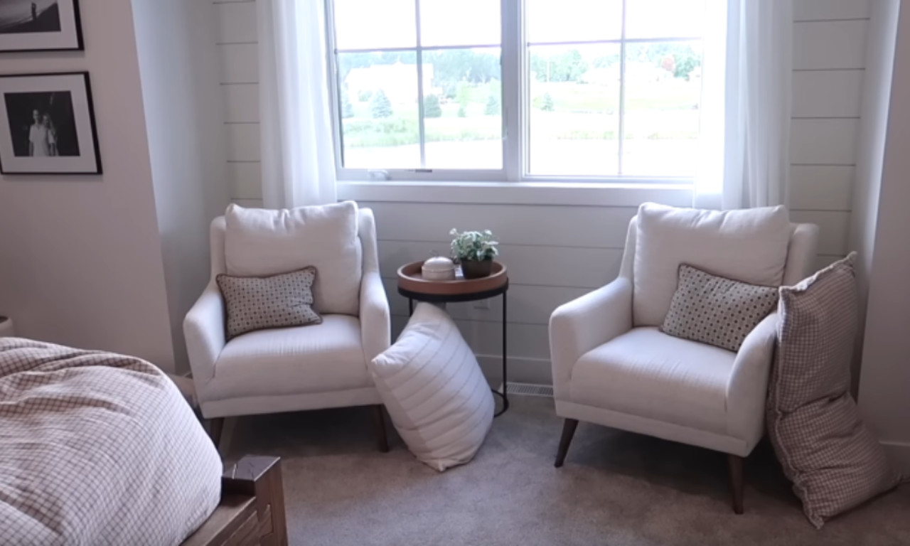A cozy bedroom reading nook featuring two matching, white upholstered armchairs flanking a small, round wooden side table. The chairs have small patterned throw pillows and are set against a backdrop of white shiplap walls and a large window with sheer curtains.