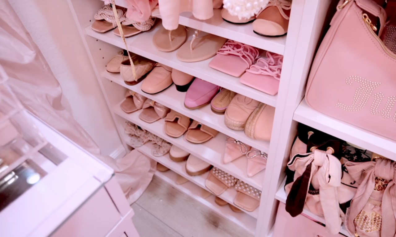 A close-up view of organized shoe shelving inside a white closet. The shelves are filled with various pairs of pink and neutral-toned flat sandals, pumps, and shoes, with a pink handbag visible on the right.