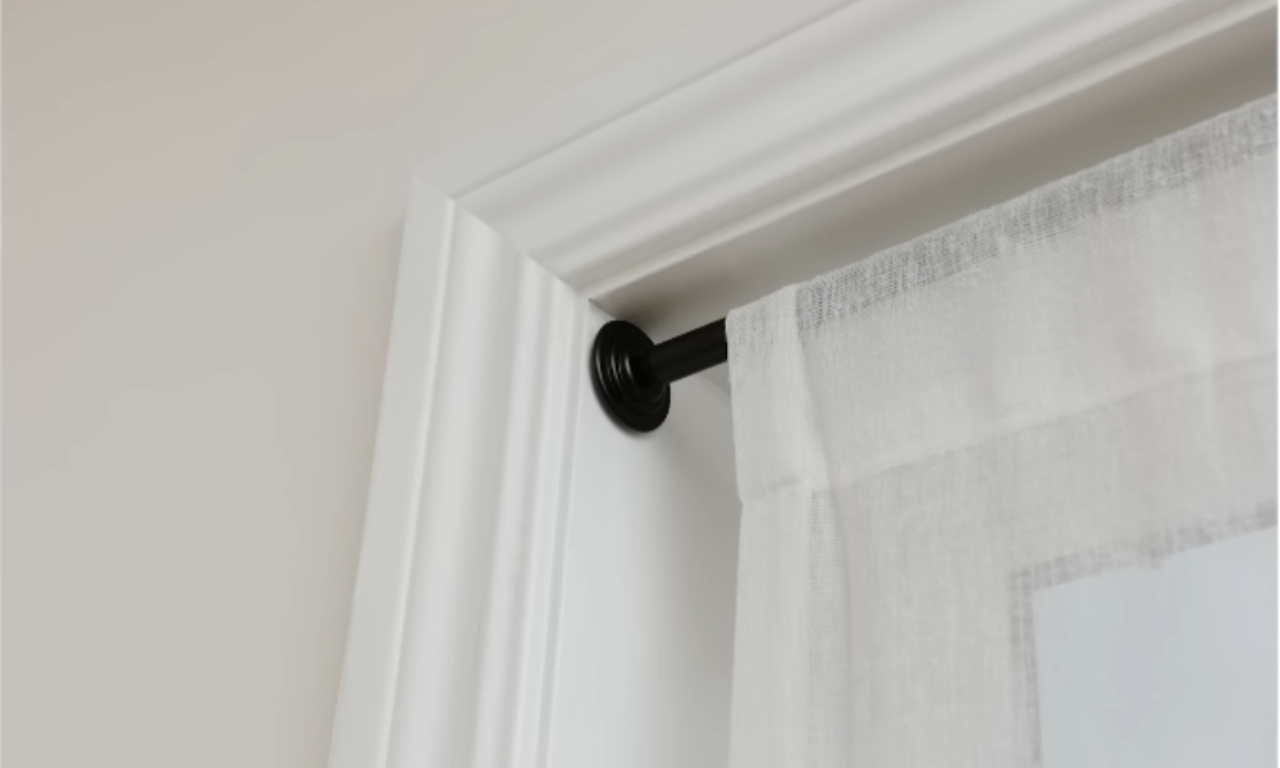 A close-up shot of a black curtain rod and white sheer curtain hung on a window frame, demonstrating a no-drill method. The rod end cap rests against the top inner corner of the white window trim.