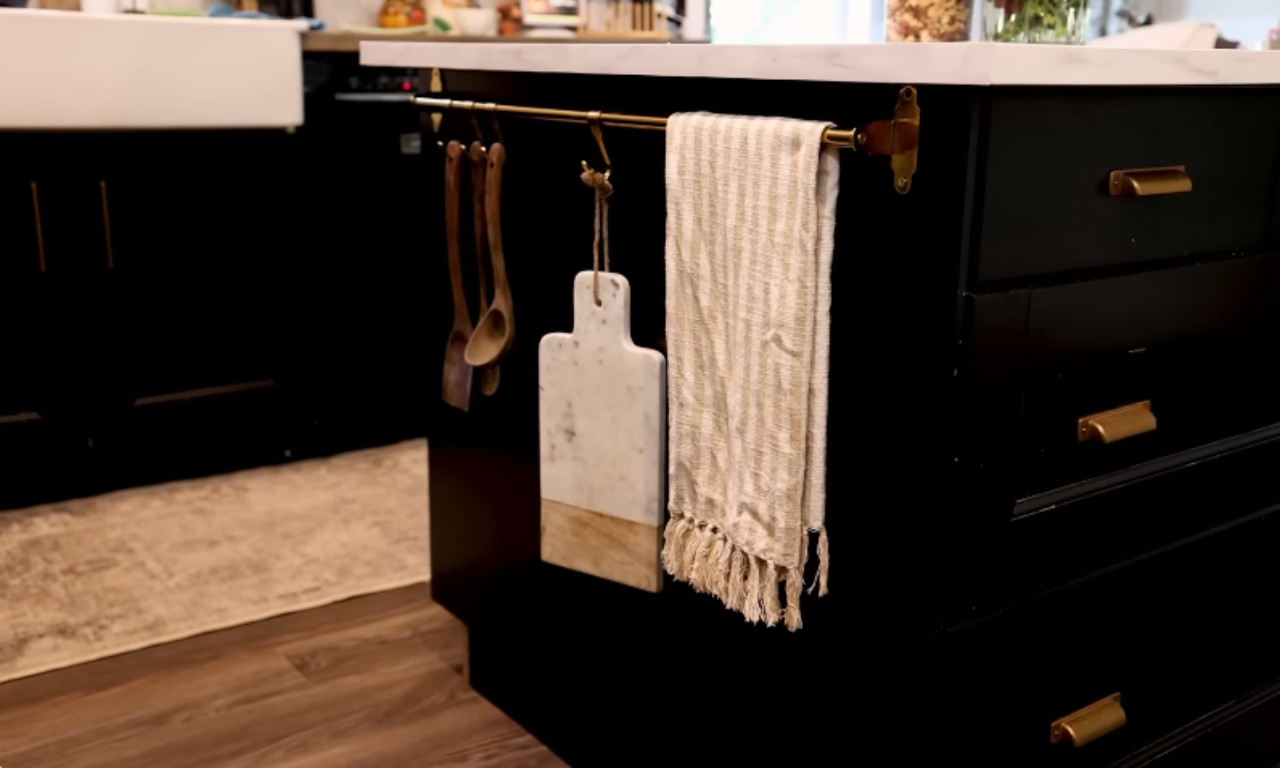 A close-up of a kitchen island customized with black cabinet fronts and gold bar handles. A brass rail is mounted to the side, holding a striped linen towel and two wooden spoons and a marble and wood cutting board suspended by hooks.