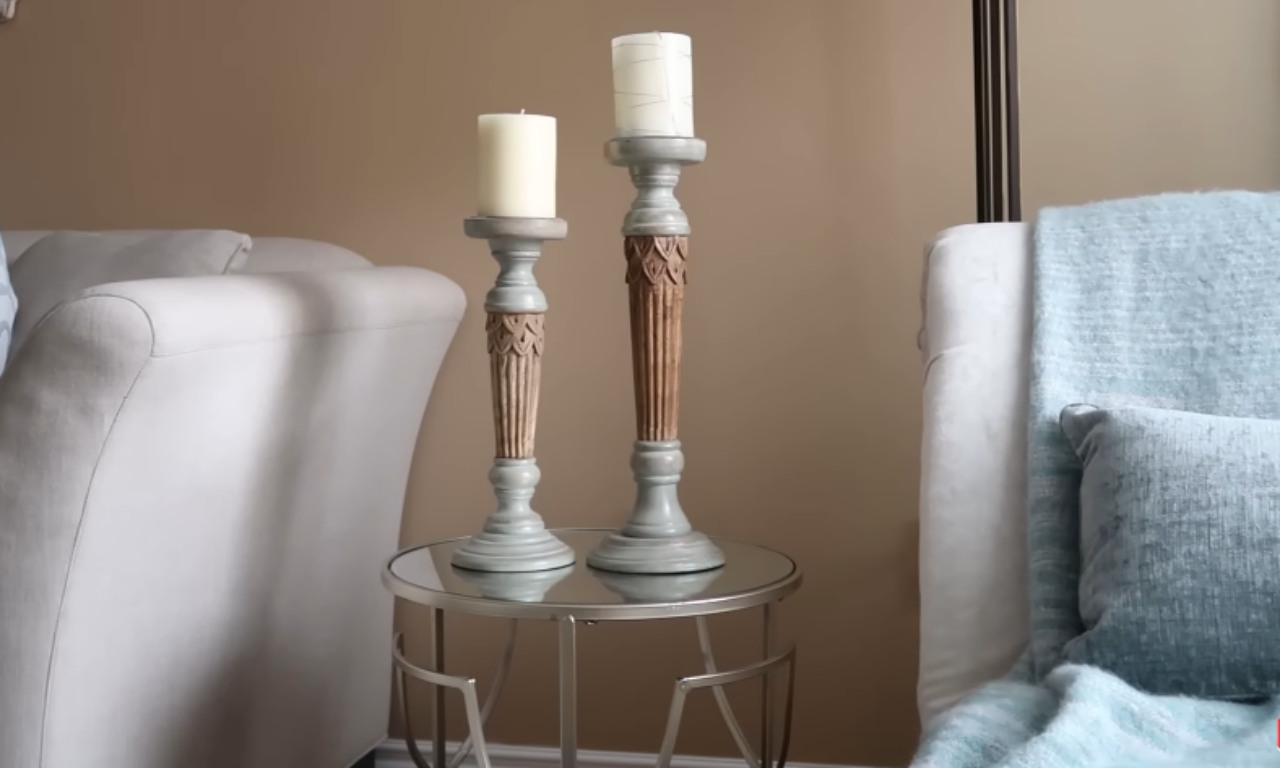 A close-up of a small, round, mirrored side table in a living room, holding two tall, decorative candle holders with white pillar candles. The candle holders have a distressed light blue base and carved wooden-look stems. A light gray sofa is visible on the left, and a white or light-colored chair/sofa with a textured light blue throw blanket and pillows is on the right.