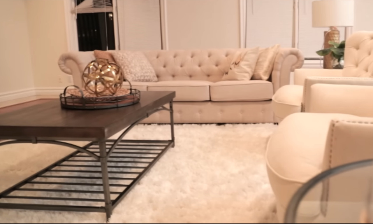 A close-up view of a chic living room corner, highlighting a beige, tufted-back sofa adorned with neutral throw pillows. The focus is on a dark wood coffee table with a decorative gold sphere on a tray, sitting on a fluffy white area rug.