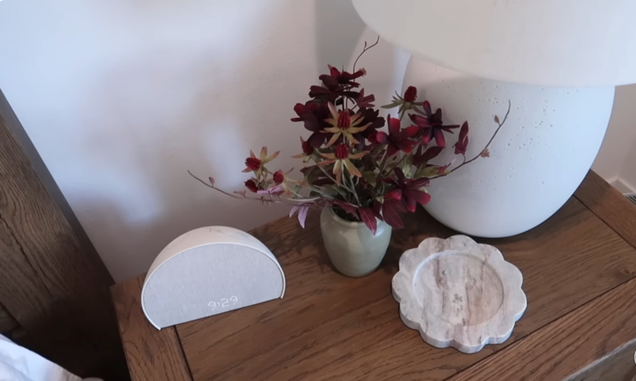 A close-up shot of a bedside table decor arrangement. The light oak table holds a modern, fabric-covered digital clock, a small green vase with a dark red and green floral arrangement, and a round, scalloped marble coaster next to a large white mushroom-shaped lamp base.