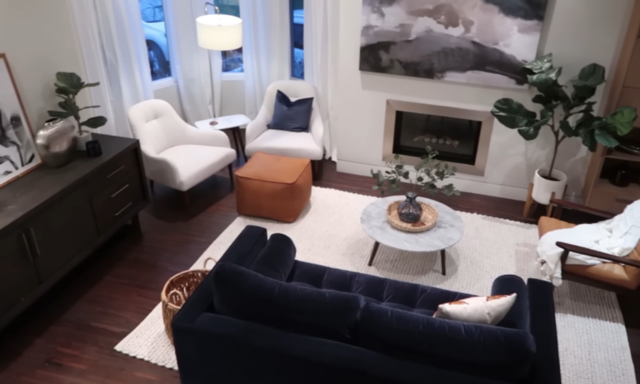 A modern, high-angle view of a living room with dark wood floors, a large navy velvet sofa, and a cream-colored textured rug anchoring the space. The room is decorated with a marble coffee table, two white armchairs, an orange leather ottoman, and several potted plants.
