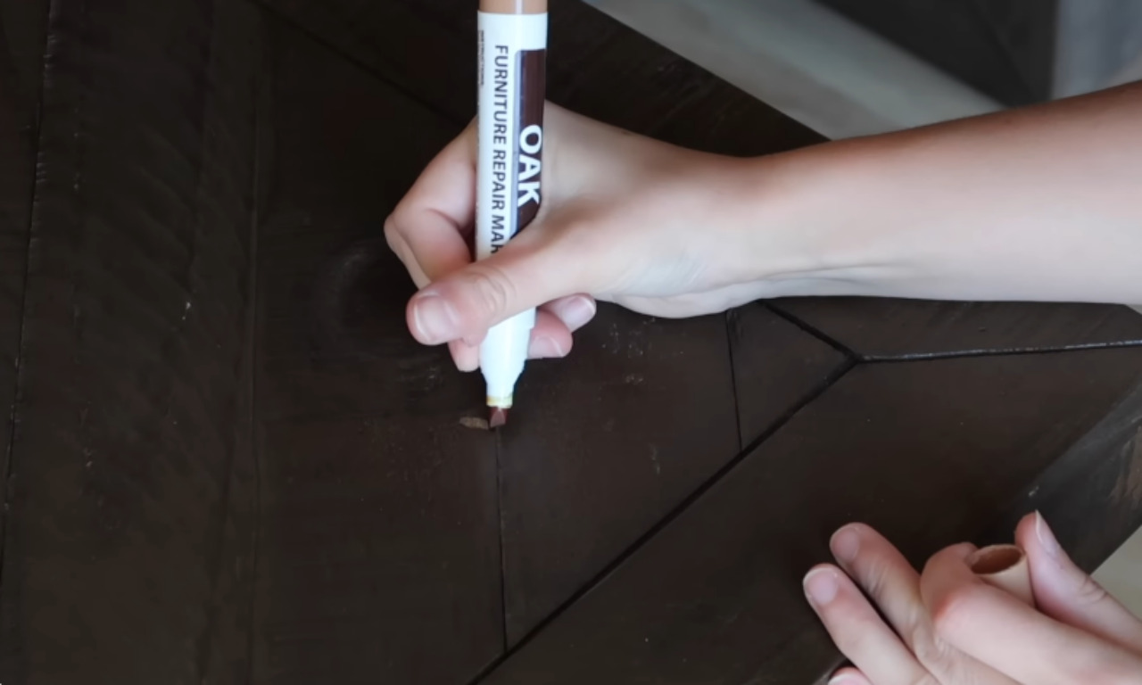 A close-up shot of a person's hand using an Oak furniture repair marker to fill in a small nick or scratch on a piece of dark brown wooden furniture. The marker tip is touching the damaged area on the tabletop, which appears to have a diagonal plank design.