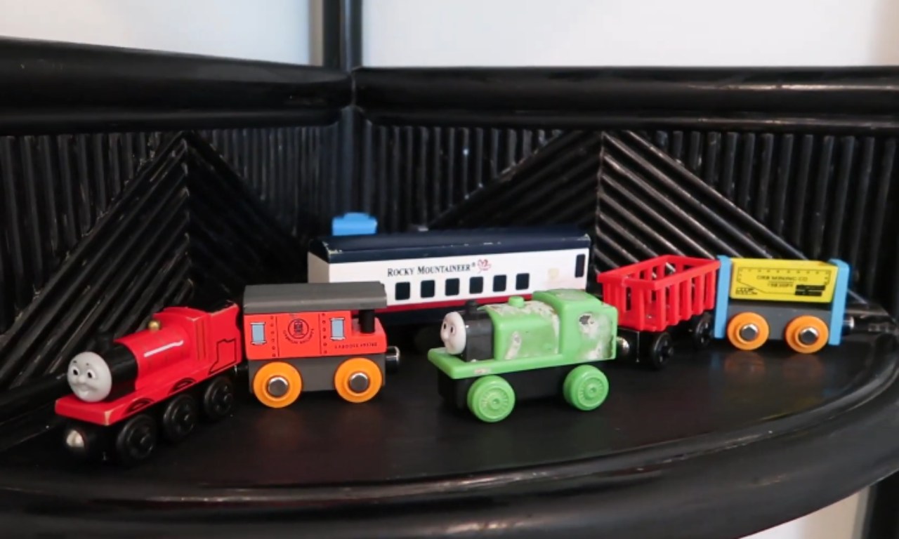 A collection of toy trains, mostly wooden railway-style, displayed on a black shelf. The trains include a red Thomas the Tank Engine, an orange boxcar, a green engine, a red hopper car, a yellow open-top car, and a longer white and blue "Rocky Mountaineer" passenger car.
