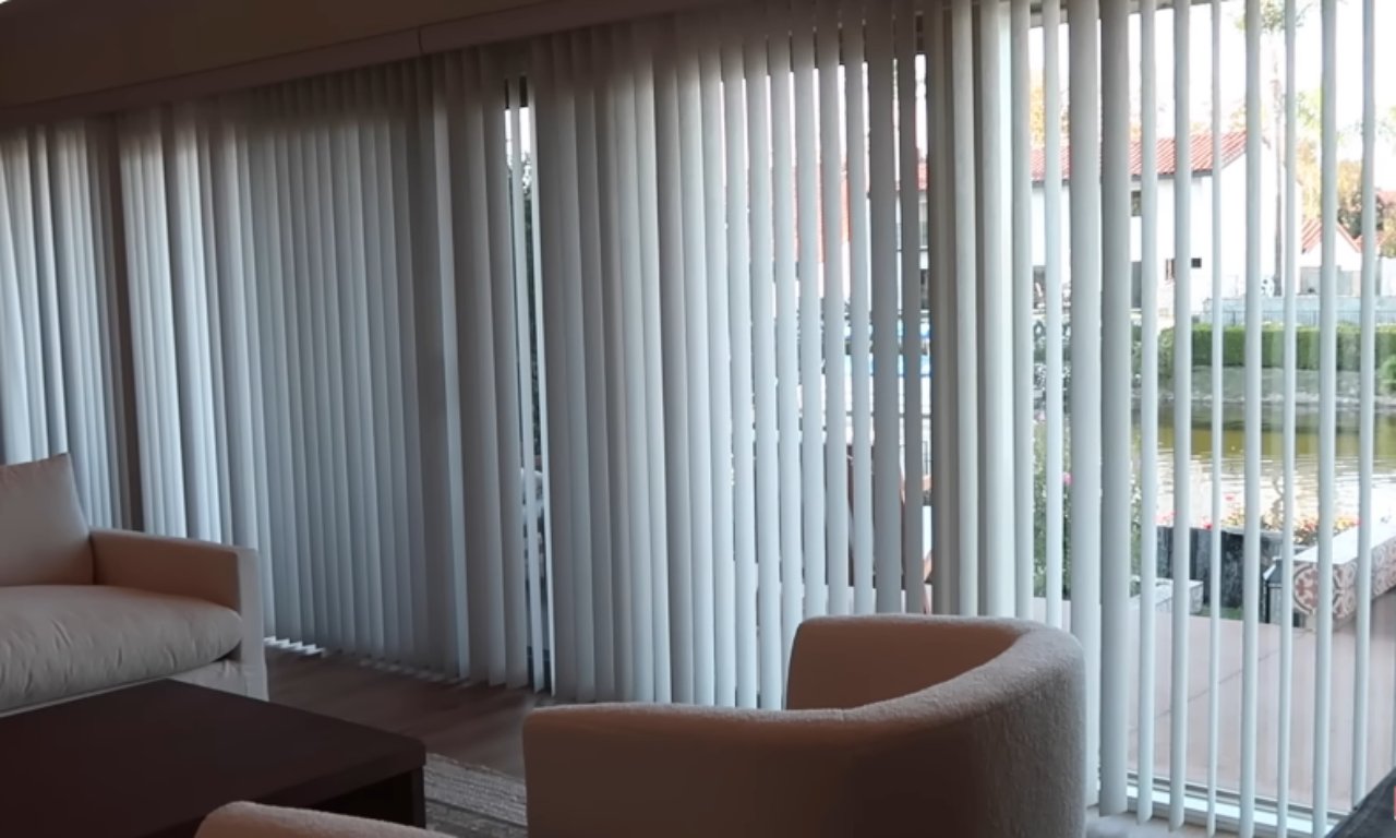 A living space with white upholstered furniture is shown, featuring a wall of vertical white blinds covering a large glass sliding door that leads out to a patio and a pond. The blinds are mostly closed, diffusing the outdoor light.