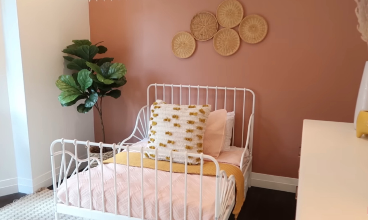 A bright, whimsical kid's bedroom featuring a white metal toddler bed with pink bedding, a yellow throw blanket, and a textured cream and yellow accent pillow. The wall behind the bed is painted a warm, muted terracotta or blush pink and decorated with a cluster of four round, woven wicker wall hangings. A tall Fiddle-Leaf Fig plant in a simple planter stands in the corner. The room's aesthetic is bohemian and soft.