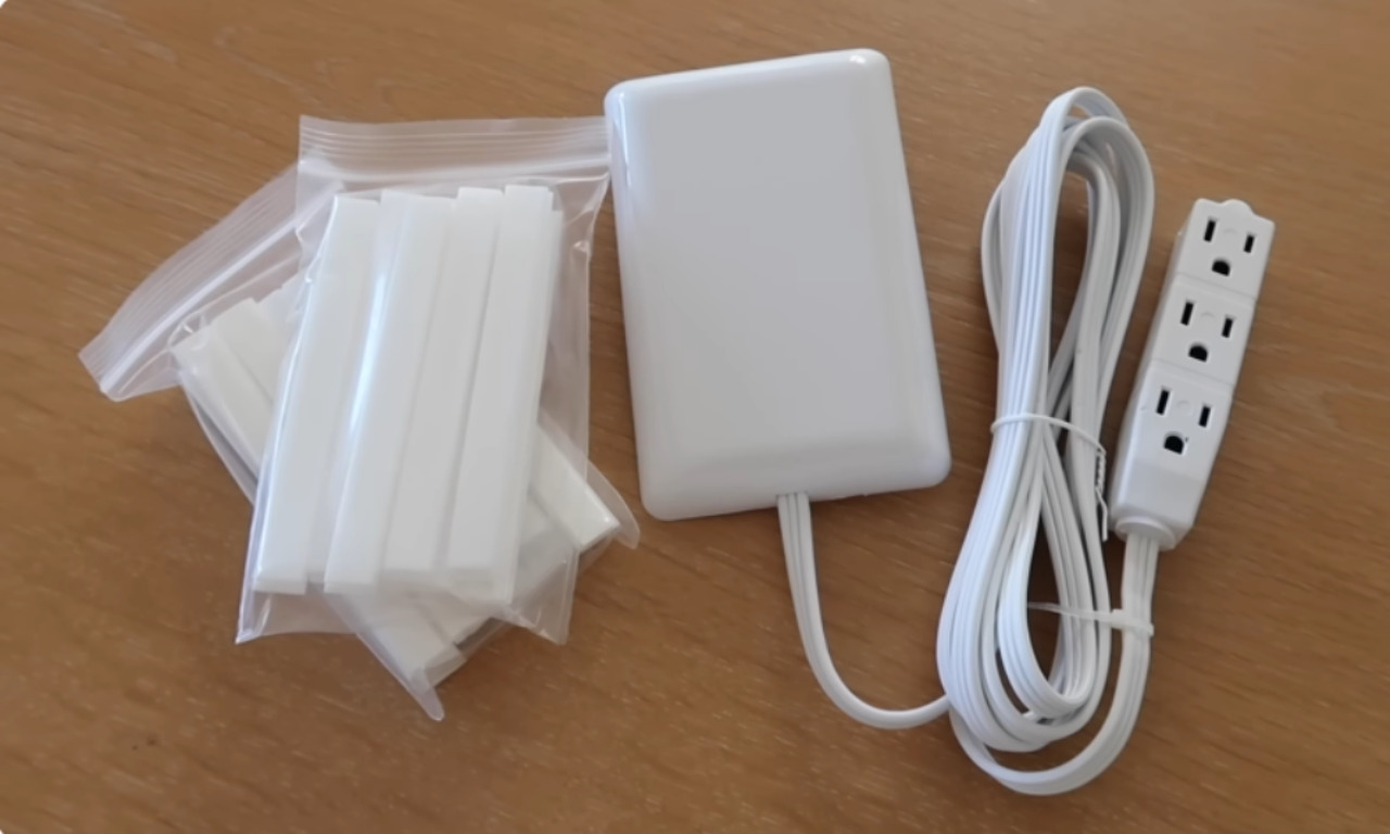 An overhead shot of three items used for cord management. On the left are several white, rectangular cable covers (or raceways) packaged in a clear plastic bag. In the center is a white, sleek surge protector or power strip with a cover. On the right is the white cord and the end of a four-outlet power strip.
