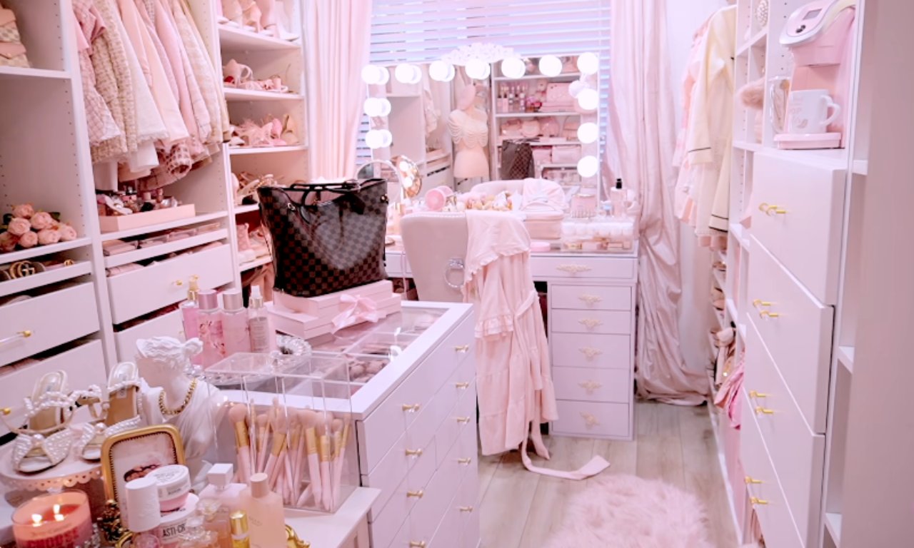 A view of a luxurious, pink-themed walk-in closet designed like a boutique. It features custom white shelving and drawers, a central vanity area with a lighted Hollywood-style mirror, and a chair. Accessories, makeup brushes, and a large handbag are displayed on the central glass-top dresser.