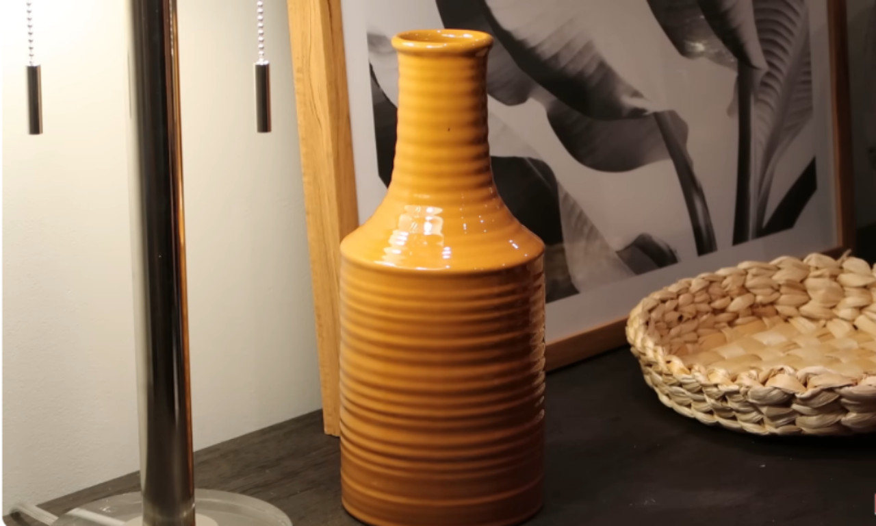 A close-up of a tabletop displaying a bright, ribbed mustard-yellow ceramic vase next to a modern silver table lamp. In the background, there is a woven basket and a framed black and white botanical print.