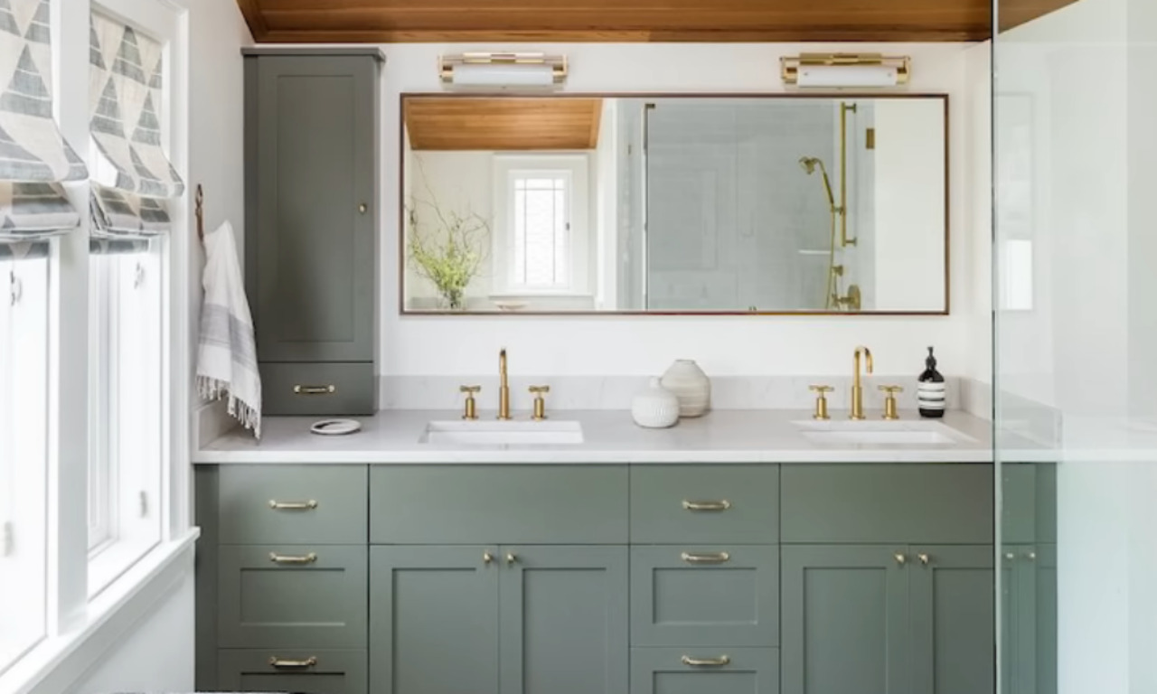 A modern bathroom features a double vanity with sage green shaker cabinets, brass hardware, and white countertops. A large, rectangular mirror framed in brass hangs above, reflecting a wood plank ceiling and a small window. Two brass and glass light fixtures are mounted above the mirror.
