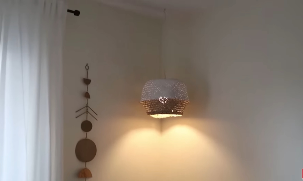A DIY pendant light hanging in the corner of a room, made from what appears to be a woven basket with a white upper section and a natural wicker or gold-toned lower section. It casts a warm glow against the light-colored walls. To the left, a piece of macrame or metal wall art with circular and semi-circular shapes hangs next to a white curtain.