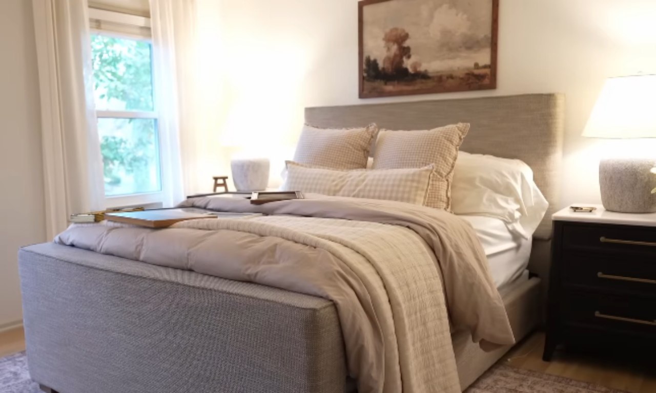 A cozy bedroom featuring a gray upholstered bed with a large headboard, covered in neutral, tan-toned bedding and several pillows. A dark nightstand with a white top is visible on the right, supporting a large table lamp with a soft white shade. A window is on the left, letting in natural light. Above the headboard, a framed vintage-style landscape painting hangs on the wall.