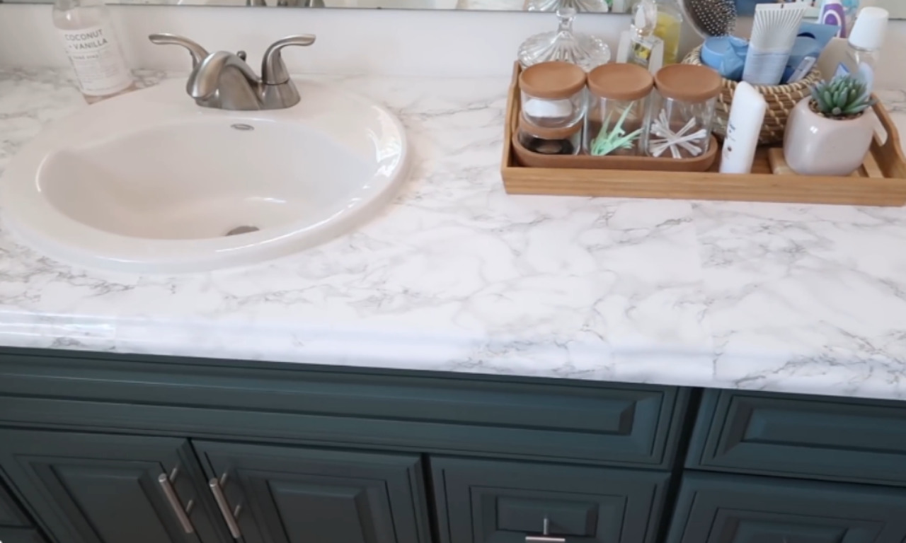 A bathroom vanity with a white countertop featuring a gray marble look and a sink. The cabinet base is painted dark teal/green. A wooden tray holds various toiletries, brushes, and small jars next to the sink.