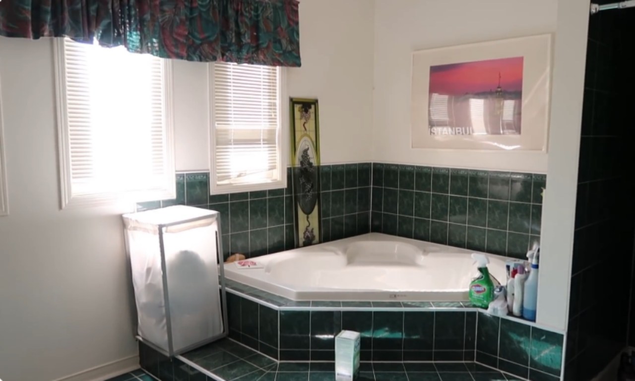 A dated bathroom with white walls and dark green, glossy, tiled walls and floor surrounding a corner bathtub. Two windows with white blinds are on the left, and a large, mesh laundry hamper sits beneath them. A framed picture of a sunset over the city of Istanbul hangs above the tub.