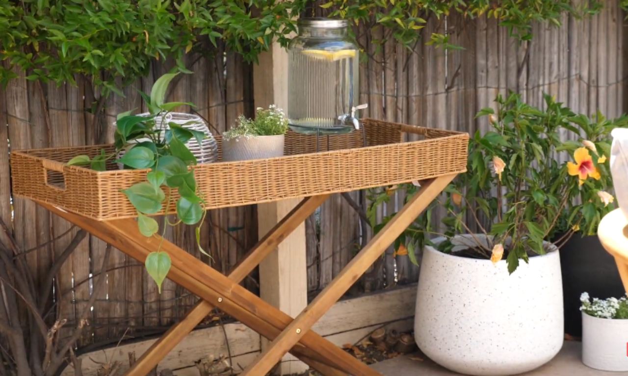 A butler's tray table with a glass drink dispenser, surrounded by potted plants including orange hibiscus and white flowers, with a bamboo fence behind.