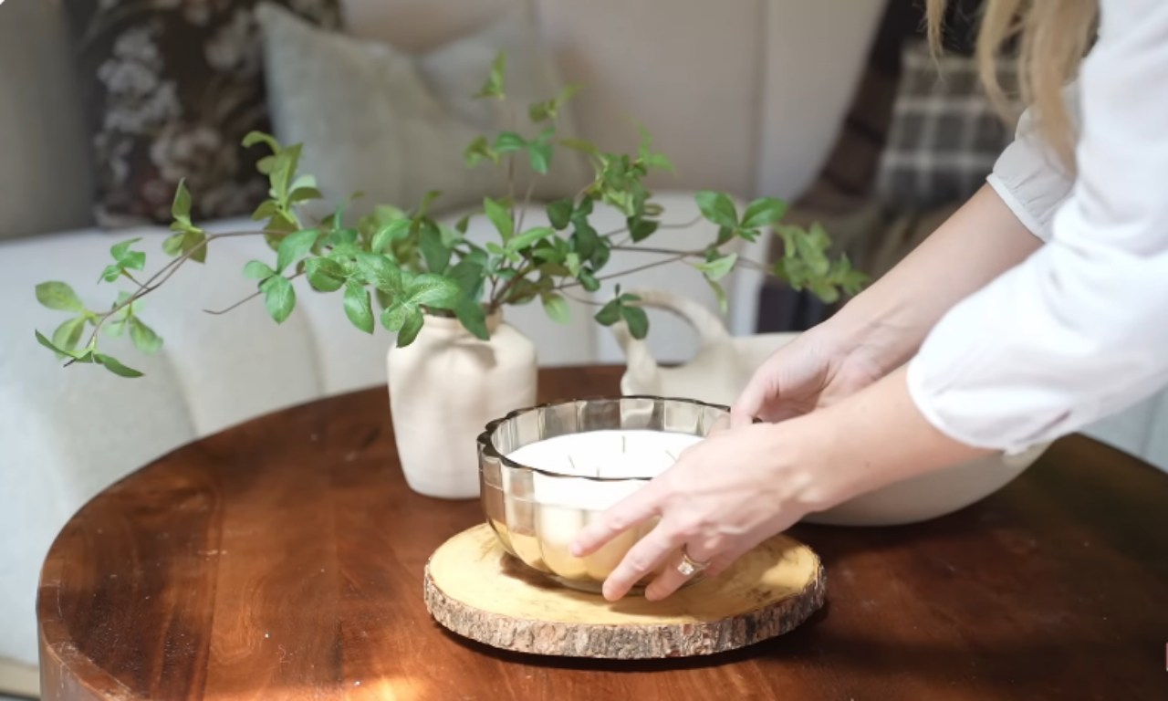 A close-up view of a large, round wooden coffee table showing a person's hands placing a wide glass bowl with a white candle on a round wood slice base. A light-colored vase with green leafy branches and a cream-colored ceramic piece are also on the table.