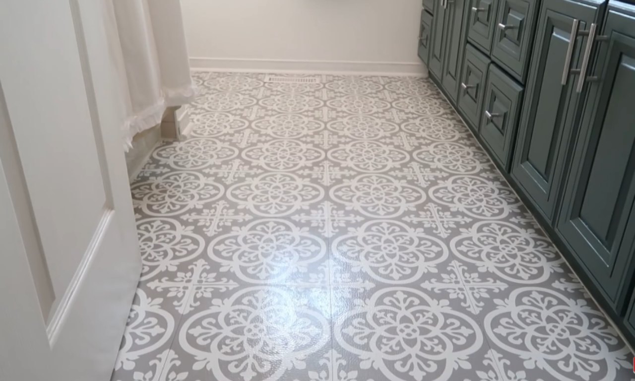 A close-up view of a bathroom floor with a new, gray and white patterned vinyl or peel-and-stick tile with a Mediterranean or floral motif. A dark teal or gray vanity is visible on the right, and a white door and shower curtain are on the left.