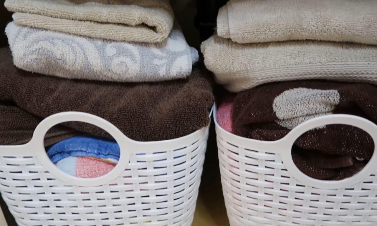 A close-up view of neatly folded, stacked towels in various shades of brown, white, and gray, stored inside two white plastic woven storage baskets.