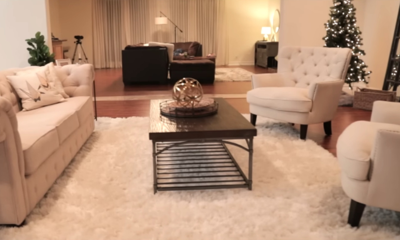 A cozy living room arranged for conversation, featuring a cream-colored tufted sofa and two matching armchairs centered around a dark wood coffee table with a metal base, all resting on a large, plush white rug. A secondary seating area with dark furniture is visible in the background, along with a lightly decorated Christmas tree.
