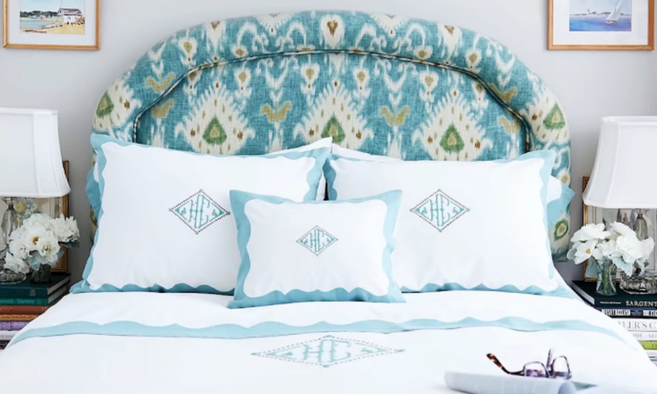 A close-up of a bed with white bedding featuring light blue trim and embroidery, including a monogram on the pillows. The bed has a striking, curved headboard with a blue and white Ikat pattern. Books and a lamp are visible on the nightstands on either side.