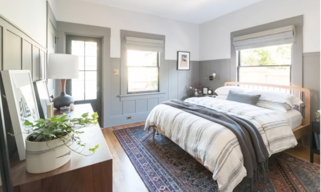 A bedroom with gray wainscoting and white walls. The room features a wooden bed frame with striped white and gray bedding and a dark gray throw blanket. A patterned area rug sits on the hardwood floor. A dark wood dresser with a potted plant is visible in the foreground, near a dark door.