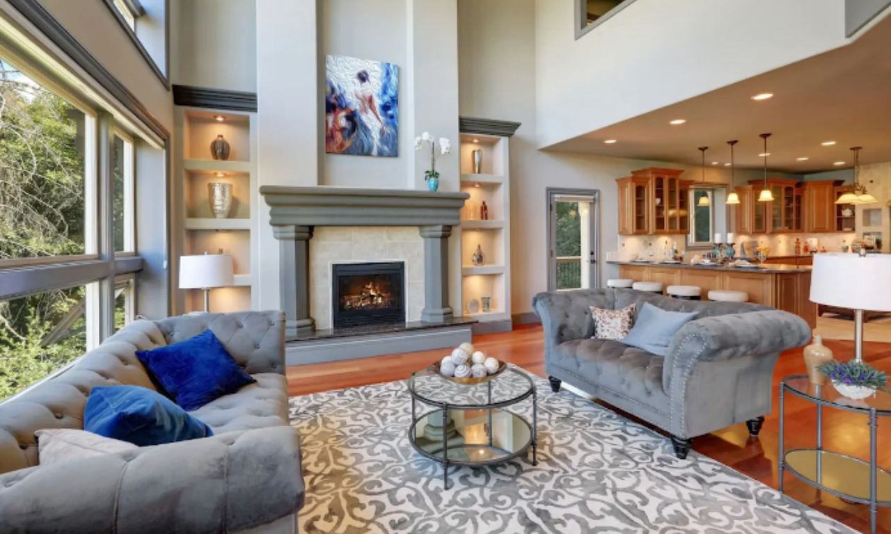 A luxurious, open-concept living area with high ceilings. The living room features a gray tufted sofa and loveseat centered on a decorative area rug and a glass coffee table. A fireplace with a prominent gray mantel is flanked by built-in vertical shelving with recessed lighting. A view of the kitchen with wooden cabinets is visible in the background.