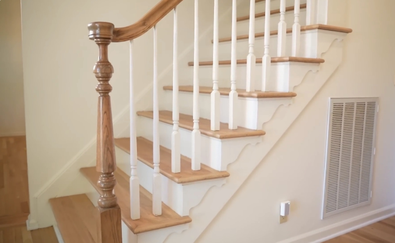 A close-up view of a traditional interior staircase. The treads and handrail are natural light wood, contrasting with the white painted risers and turned balusters. The stringer features decorative scalloped trim details. A large, ornate wooden newel post is visible at the bottom.