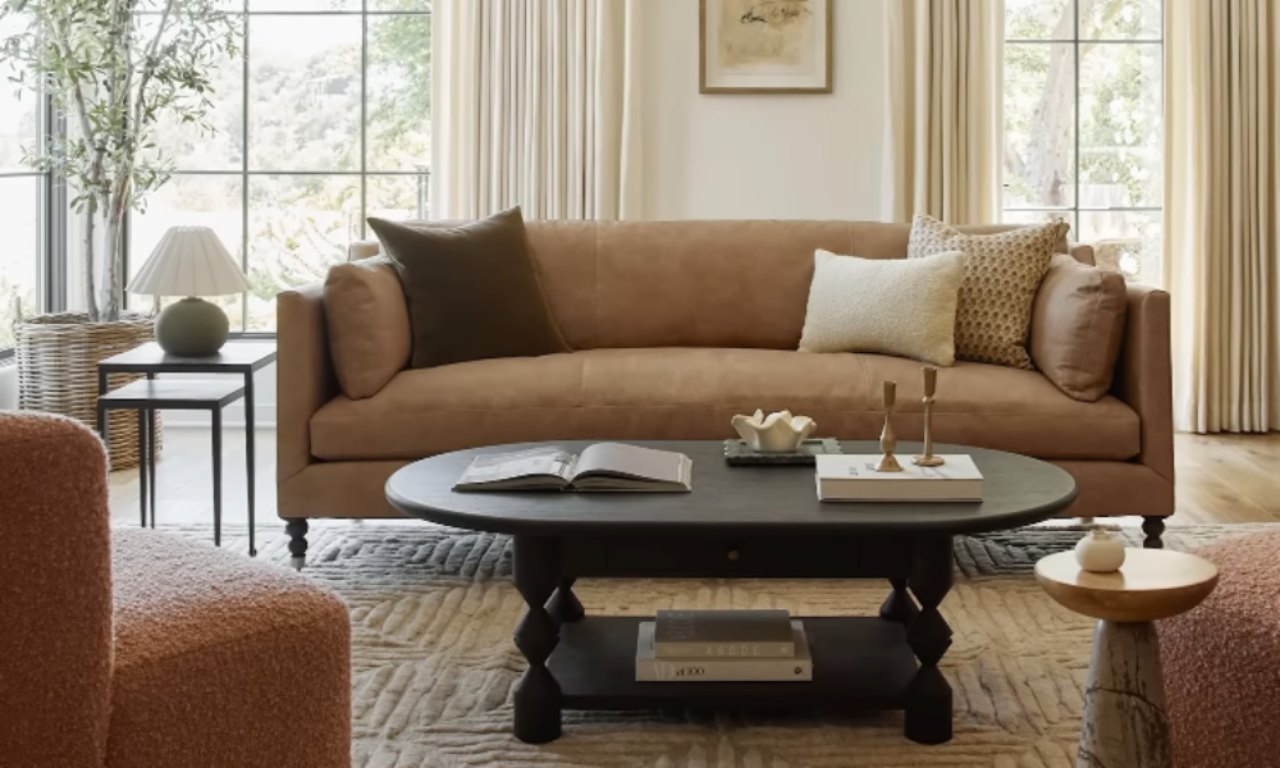 A cozy, modern living room featuring a tan leather sofa with several throw pillows, a dark oval coffee table in front of it, and a pair of pinkish-brown textured accent chairs framing the view. Large windows with sheer curtains let in natural light.